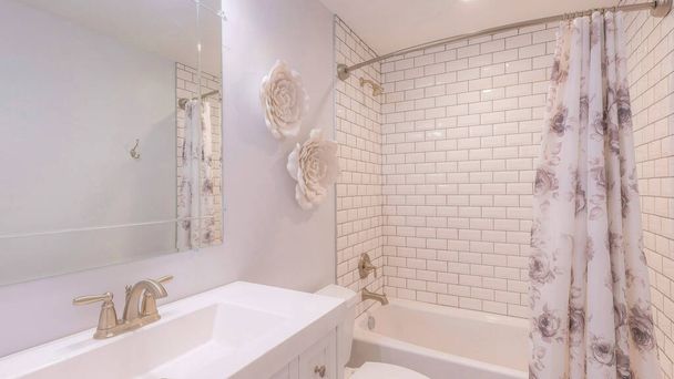 Panorama Salle de bain traditionnelle avec thème floral blanc et baignoire douche avec carreaux de métro entourent. Il y a un lavabo avec dessus solide blanc et miroir près des toilettes avec deux fleurs blanches sur le mur. - Photo, image