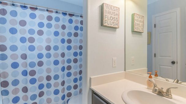 Πανόραμα Εσωτερικό μπάνιο με νιπτήρα ματαιοδοξίας και κουρτίνα ντους με τυπωμένες πουά. Υπάρχει μια τουαλέτα κοντά στο νεροχύτη ματαιοδοξία με διακόσμηση δήλωση στον τοίχο και τον καθρέφτη με μια αντανάκλαση των δύο άσπρες πόρτες. - Φωτογραφία, εικόνα