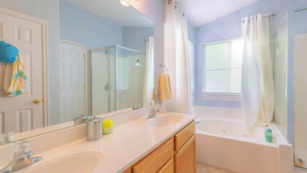 Panorama Sun flare Cuarto de baño con paredes de color azul claro y ventanas en la esquina con cortinas. Hay lavabos de tocador doble con espejo cerca de la bañera de la esquina al lado del puesto de ducha a la izquierda. - Foto, Imagen