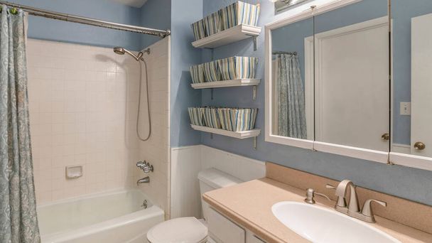 Panorama Wnętrze łazienki z koszami na pościel na pływających półkach. Po lewej stronie znajduje się zlewozmywak z lustrzanymi szafkami na ścianie przy toalecie obok wanny z płytkami ściennymi.. - Zdjęcie, obraz