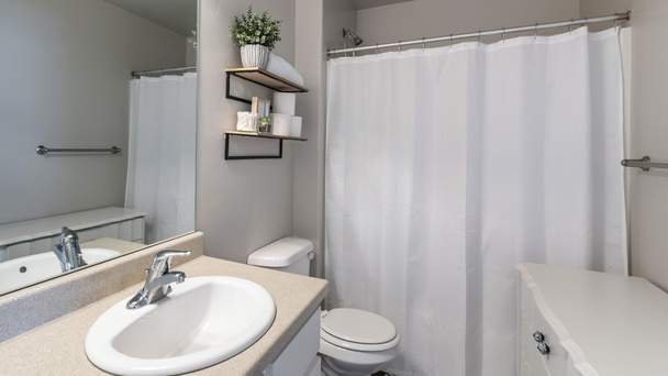 白い床キャビネットと虚栄心シンク付きのバスルームのパノラマインテリア。白いシャワーカーテンの近くの棚に植物、タオル、トイレットペーパーとトイレの近くの虚栄心の上に鏡があります。. - 写真・画像