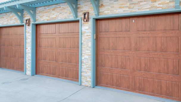 Panorama Garage exterior with wall-mounted light blue pergola and doorframe. There are three wooden sectional doors and stone veneer wall sidings. - Photo, Image