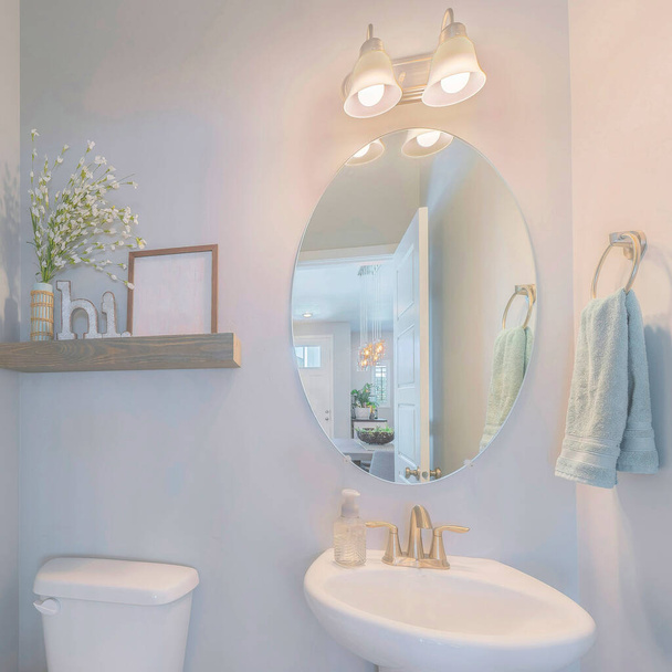 Square Powder room interior with decorations on the shelf. There is a wall mount toilet paper holder on the left near the toilet bowl beside the pedestal sink with oval mirror and hanging towel. - Photo, Image
