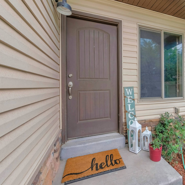Square Facade of house with beige vinyl lap siding and gray front door near the window. Entrance of a home with welcome signboard and white lanterns on the concrete doorstep near the plants on the right. - Photo, Image