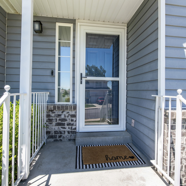 Quadratische Veranda eines Hauses mit gläserner Sturmtür mit Seitentür. Eingang eines Hauses mit Türklingel und Fußmatte an der Haustür mit Geländer und einer Außenseite aus grauem Vinyl und Ziegelsteinen. - Foto, Bild