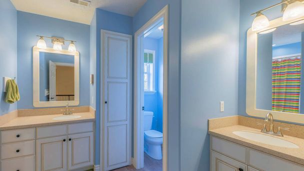 Ванная комната Panorama Master со светло-голубым интерьером и двумя раковинами. Существует туалетная комната между двумя раковинами тщеславия с зелеными коврами и цветной полосатой занавеской для душа справа. - Фото, изображение
