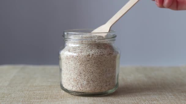 Psyllium is a plantain husk. A glass jar with a wooden spoon from which Psyllium sprinkles on a beige and gray background - Footage, Video