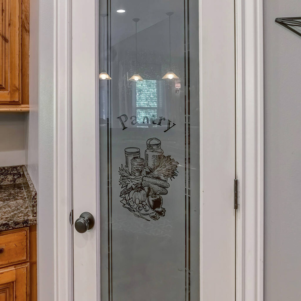 Kwadratowe drzwi spiżarni z ozdobnym szklanym panelem z oznakowaniem spiżarni. Drzwi spiżarni wewnątrz kuchni z drewnianymi szafkami i brązowym granitowym blatem. - Zdjęcie, obraz