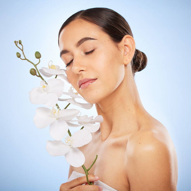 Schoonheid, huidverzorging en vrouw met een orchidee in een studio voor een gezondheid, wellness en natuurlijk gezicht routine. Cosmetische, zelfverzorging en meisje model met heldere huid van spa gezichtsbehandeling door blauwe achtergrond - Foto, afbeelding