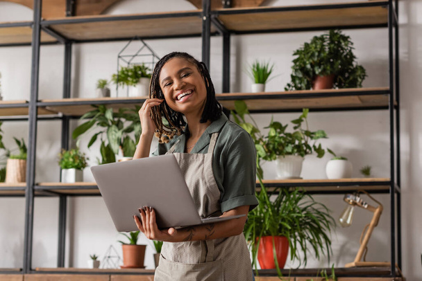 cheerful african american florist with dreadlocks standing with laptop near rack with green potted plants on blurred background - Foto, Imagem