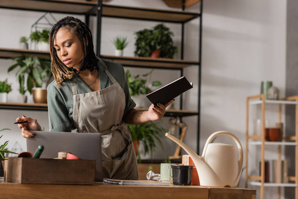 stylish african american woman in apron holding notebook and looking in box with flowerpots near laptop - Foto, afbeelding