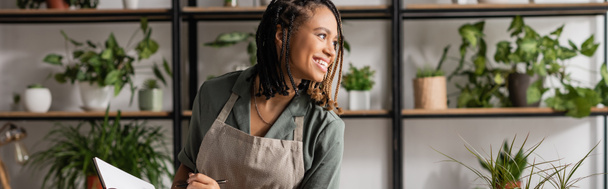 cheerful african american florist holding notebook and looking away near rack with plants on blurred background, banner - Foto, Imagem