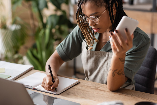 молодий афроамериканський флорист в окулярах тримає смартфон і пише в блокноті біля розмитого ноутбука в квітковому магазині
 - Фото, зображення