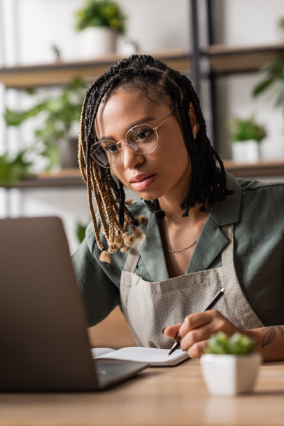 focalizzato fiorista afroamericano in occhiali da vista guardando laptop e scrittura ordine nel negozio di fiori - Foto, immagini