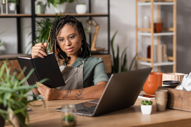 stílusos afro-amerikai virágárus gazdaság notebook, miközben ül a munkahelyen közelében laptop virágüzlet - Fotó, kép