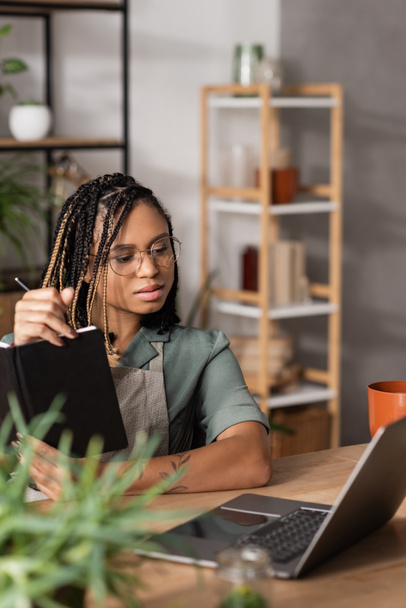 jovem florista afro-americano em óculos segurando notebook e olhando para o laptop enquanto trabalhava na loja de flores - Foto, Imagem