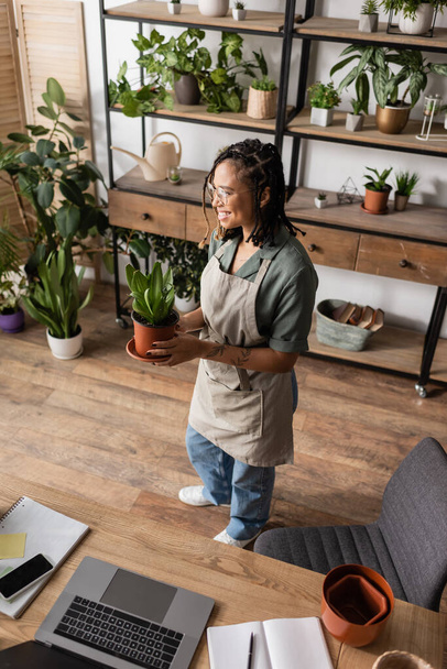 vista ad alto angolo di felice fiorista afroamericano che tiene pianta in vaso vicino alla scrivania con dispositivi nel negozio di fiori - Foto, immagini