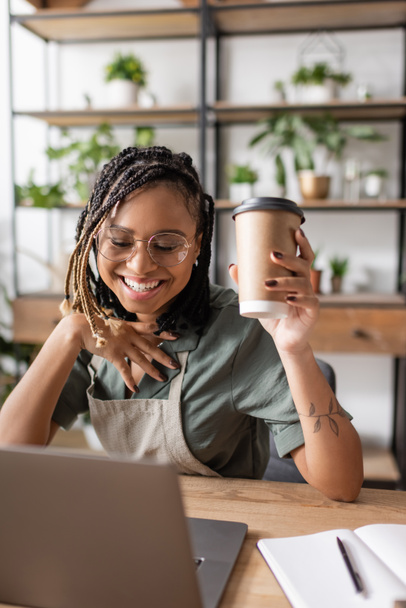 フラワーショップでのビデオ通話中にノートパソコンの近くで笑顔になるコーヒーを持つトレンディーなアフリカ系アメリカ人花屋 - 写真・画像