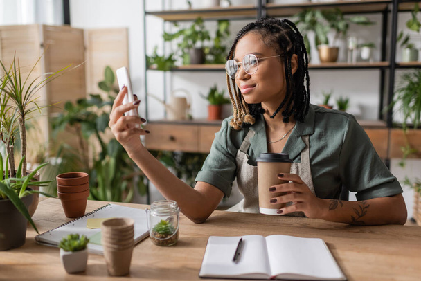 усміхнений афроамериканський флорист з паперовою чашкою дивиться на мобільний телефон біля порожнього блокнота і вазони в магазині
 - Фото, зображення