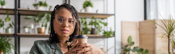 floristería afroamericana en gafas con estilo y rastas sosteniendo café para ir y mirando a la cámara en la tienda de flores, pancarta - Foto, imagen