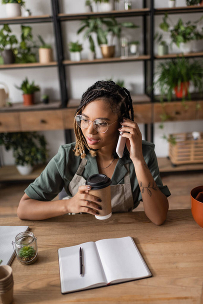 young african american florist in apron and eyeglasses holding takeaway drink and talking on smartphone in flower shop - Photo, Image