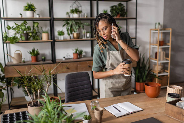 Kağıt bardaklı Afrikalı Amerikalı çiçekçi cep telefonuyla konuşuyor ve saksı bitkilerinin yanındaki boş defterlere bakıyor. - Fotoğraf, Görsel
