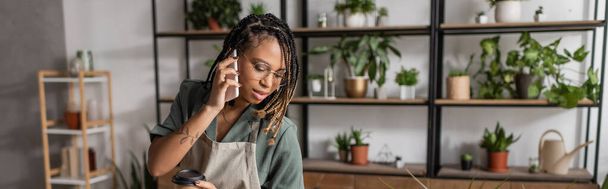 young african american florist in eyeglasses talking on mobile phone near potted plants on blurred background, banner - Photo, Image