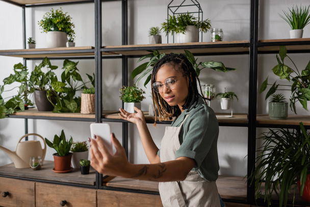 νεαρός αφροαμερικανός ανθοπώλης με κινητό τηλέφωνο δείχνει σε ποικιλία φυτών στο ανθοπωλείο - Φωτογραφία, εικόνα
