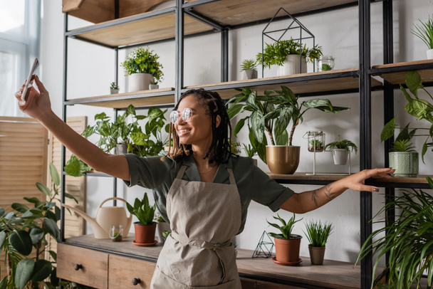 sourire afro-américain fleuriste avec téléphone portable debout près de rack avec des plantes en pot lors de la commande en ligne - Photo, image