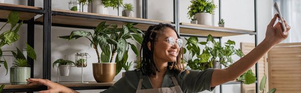 smiling african american florist in eyeglasses showing assortment of potted plants during video call on smartphone, banner - Фото, изображение