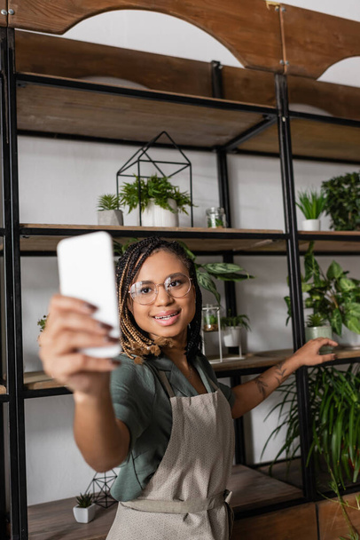nuori ja tyylikäs afrikkalainen amerikkalainen kukkakauppa, jolla on hämärtynyt älypuhelin samalla kun näytetään teline, jossa on valikoima kasveja videopuhelun aikana - Valokuva, kuva
