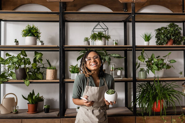 happy african american florist with mobile phone looking at camera near rack with different potted plants - Photo, Image