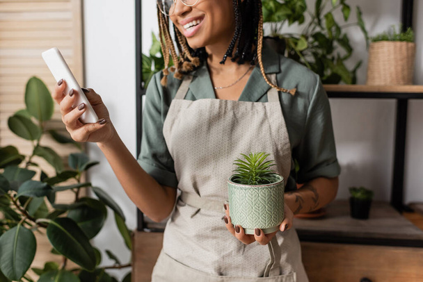 przycięty widok uśmiechniętej afrykańskiej kwiaciarni amerykańskiej z roślin doniczkowych gospodarstwa telefon komórkowy podczas zamówienia online - Zdjęcie, obraz