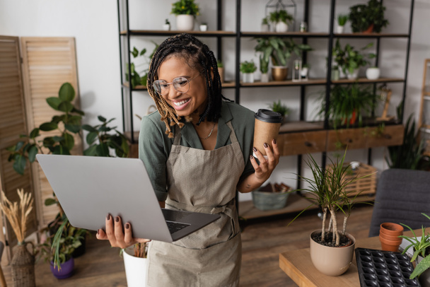 joyful african american florist in eyeglasses and apron holding coffee to go during video call in flower shop - Photo, Image