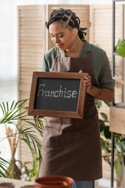 young and trendy african american florist in apron holding board with franchise lettering in flower shop - Photo, Image