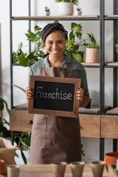 cheerful african american florist with trendy hairstyle holding board with franchise lettering near rack with plants - Photo, Image