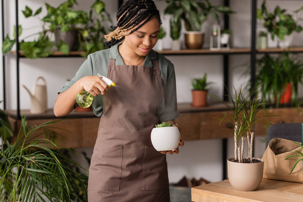 african american florist with dreadlocks spraying water on potted plant in flower shop - Zdjęcie, obraz