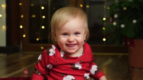 Una bella ragazza bionda di un anno con un vestito natalizio si siede e sorride guardando la macchina fotografica, ghirlanda sullo sfondo. Bambini e vacanze di Capodanno - Filmati, video