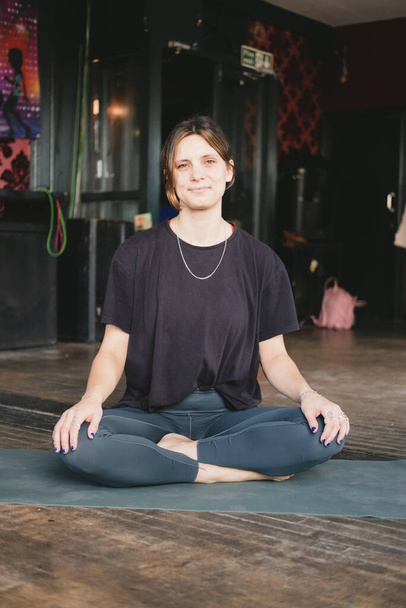 Foto vertical de uma nova professora de ioga feminino sorridente descansando em pose fácil (sukhasana) durante sua prática de ioga de fluxo de vinyasa vestindo roupas esportivas - Foto, Imagem