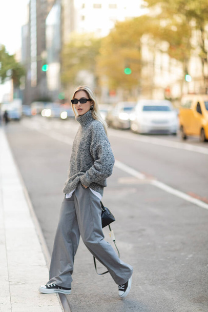 full length of stylish woman in sunglasses and grey outfit holding handbag while walking on street of New York city  - 写真・画像