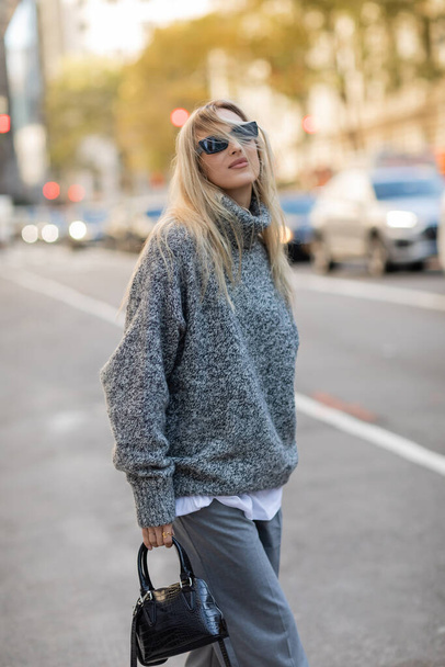 potěšená žena v slunečních brýlích a módní oblečení drží kabelku, zatímco stojí na ulici New York City  - Fotografie, Obrázek
