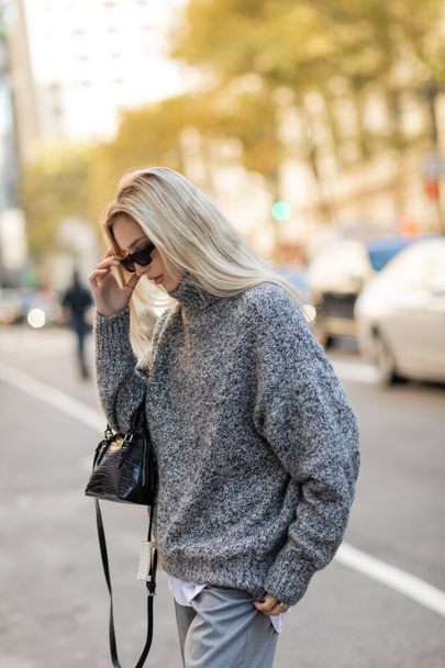 blonde woman in grey sweater holding handbag and adjusting trendy sunglasses on street of New York city  - Фото, зображення