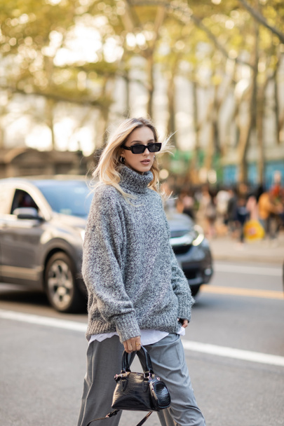 trendy woman in grey sweater and sunglasses holding handbag while walking on street of New York city  - Foto, Imagen