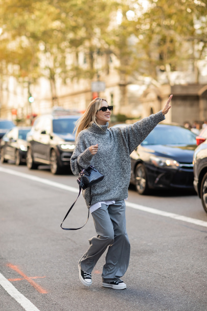 full length of cheerful woman in sunglasses and grey outfit holding handbag and catching cab on street of New York - Photo, Image