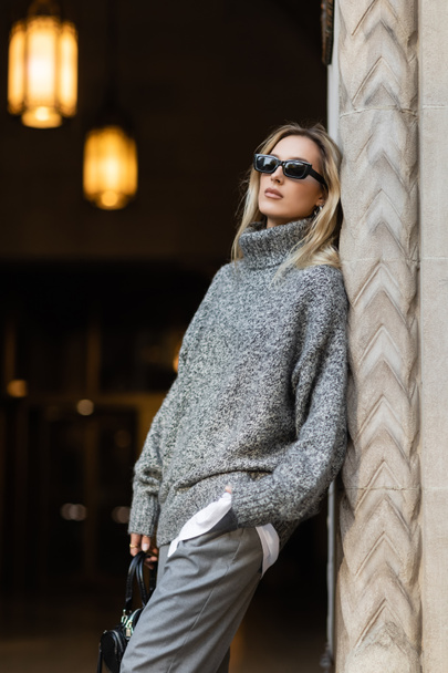 Blondine mit Sonnenbrille und grauem Outfit hält Handtasche, während sie mit der Hand in der Tasche in der Nähe eines Gebäudes in New York steht  - Foto, Bild
