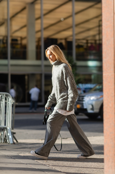 full length of stylish young woman with blonde hair walking in trendy winter outfit in New York  - Zdjęcie, obraz