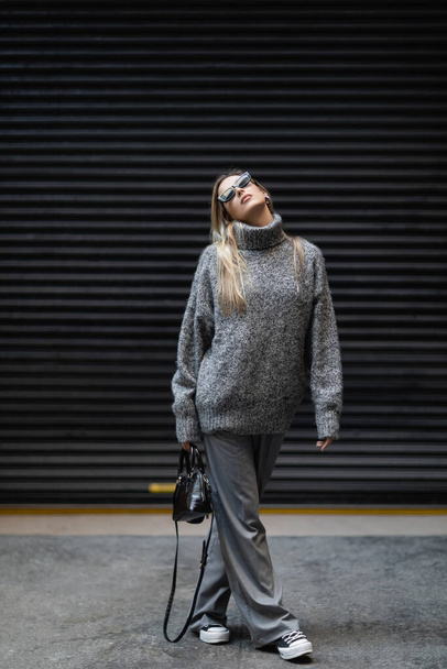 longitud completa de mujer rubia en suéter de invierno y gafas de sol caminando con bolso en la calle urbana  - Foto, Imagen