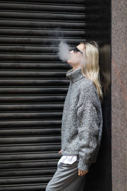 Seitenansicht einer jungen Frau in Winterpullover und Sonnenbrille, die E-Zigarette raucht, während sie auf der Straße steht  - Foto, Bild