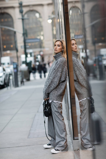 full length of blonde woman in winter sweater standing with handbag on urban street in New York  - Foto, immagini