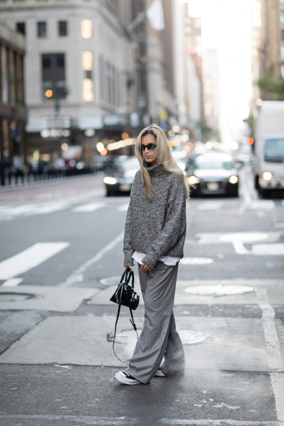 full length of blonde woman in knitted sweater and sunglasses walking with trendy handbag on urban street in New York  - Photo, image